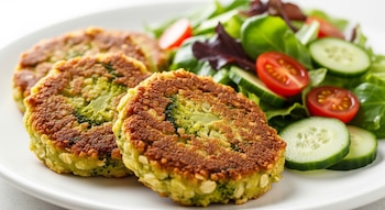 Primer plano de tres tortitas doradas de brócoli y avena en un plato blanco, junto a una ensalada fresca con lechuga, pepino en rodajas y tomates cherry partidos por la mitad.