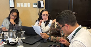 Cuatro personas sentadas alrededor de una mesa de trabajo con computadoras portátiles, documentos, vasos y teléfonos móviles