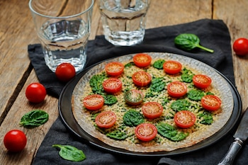Pizza fina con espinacas y mitades de tomate cherry sobre un plato negro, servida junto a dos vasos de agua sobre una mesa de madera.
