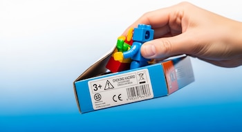 Una mano sostiene una caja abierta de juguete con figuras de bloques. La caja muestra el símbolo 3+, una advertencia de peligro de asfixia, marca CE y código de barras.