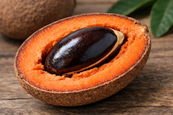 Mamey cortado por la mitad muestra pulpa naranja y semilla oscura. Otro mamey y hojas verdes son visibles al fondo sobre una superficie de madera.