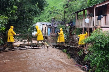 Personas con impermeables amarillos cruzan un puente de madera sobre un río caudaloso y trasladan bolsas desde una vivienda