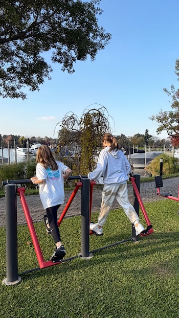 Vista trasera de dos niñas utilizando aparatos de ejercicio al aire libre en un parque, con un río y barcos al fondo bajo un cielo azul claro
