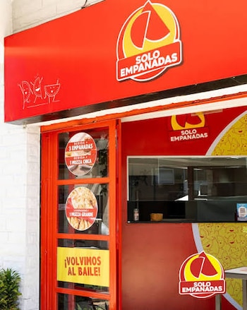 Fachada roja de un local "Solo Empanadas" con logo, puerta roja con tres pegatinas circulares y un cartel amarillo, y una ventana que muestra el interior