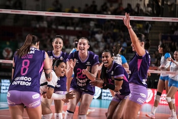 Jugadoras de voleibol con uniforme morado sonríen y celebran en la cancha cerca de la red ante los espectadores