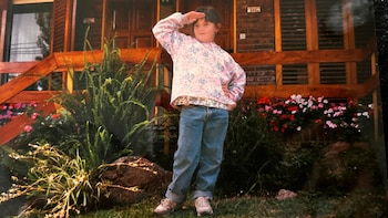Niña Laura Scianca de pie frente a una casa de ladrillo y madera, con plantas verdes y flores, su mano derecha sobre la frente mirando al frente
