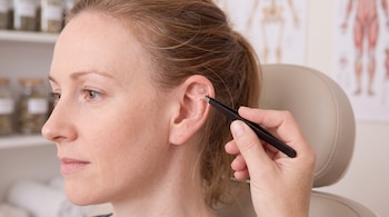 Mujer de perfil con el cabello recogido. Una mano sostiene pinzas negras y coloca pequeñas esferas doradas en su oreja. Estanterías y gráfico anatómico al fondo.