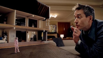 Matthew Macfadyen, con traje oscuro, observa atentamente una pequeña figura de mujer en pijama rosa, parada junto a una casa de muñecas de dos pisos