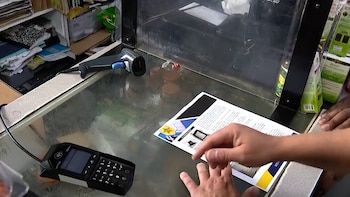 Primer plano de un mostrador con un terminal móvil POS, un escáner de códigos de barras, manos de dos personas y un documento impreso