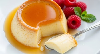 Un flan de yogur con caramelo derretido, una porción en una cuchara, y decorado con frambuesas rojas y hojas de menta verde en un plato blanco.