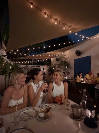 Tres mujeres sentadas a una mesa de restaurante al aire libre por la noche, con luces de guirnalda colgando y toldos claros. Hay bebidas en copas sobre la mesa