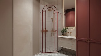 Baño moderno con ducha esquinera de marco rojizo y vidrio acanalado, lavabo flotante con grifería dorada y gabinetes color borgoña. Espejo iluminado.
