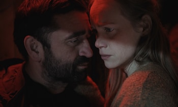 Primer plano de un hombre con barba y una mujer con cabello rubio mirándose fijamente, iluminados por una luz rojiza en un ambiente oscuro