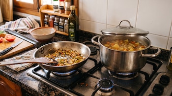 Una sartén con verduras sofritas y una olla de estofado sobre fogones de gas encendidos, en una mesa de trabajo con frascos de especias.