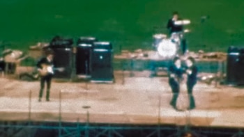 Cuatro músicos actúan sobre un escenario elevado con amplificadores y una batería ante un fondo de césped verde