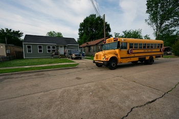 Un autobús escolar amarillo con "Caddo Parish Schools" en el lateral está estacionado en una calle de hormigón. Detrás, una casa verde y un coche gris bajo un cielo con nubes