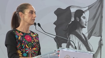 Claudia Sheinbaum habla ante dos micrófonos en un podio frente a un mural con una mujer y la bandera mexicana.