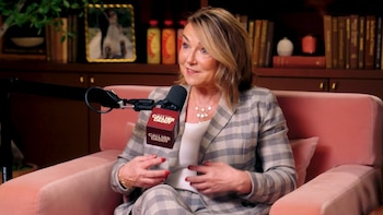 Mujer de mediana edad con cabello rubio y traje a cuadros gris y blanco sentada en un sillón rosa, hablando a un micrófono con el logo de Call Her Daddy