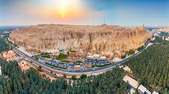 Vista aérea de una montaña rocosa con grietas, un edificio con estacionamiento de automóviles y un extenso palmeral bajo el sol.