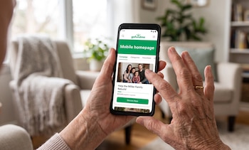 Manos sostienen un teléfono móvil negro que muestra la aplicación GoFundMe con una foto familiar y un botón de donación. Fondo de sala difuminado