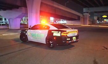 Coche de policía blanco y negro con luces intermitentes bajo un paso elevado de hormigón de noche. Hay cinta policial amarilla