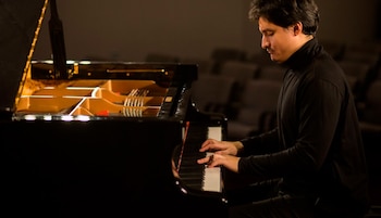 Un hombre toca un piano de cola en un escenario poco iluminado, visto de perfil con las manos sobre las teclas y la tapa del piano levantada
