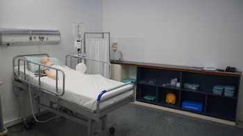 Un maniquí médico yace en una cama de hospital junto a un pie de suero, un lavabo y muebles con insumos de salud