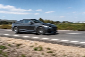 Vista lateral de un coche gris en movimiento sobre el asfalto de una carretera de dos carriles con vegetación al fondo bajo un cielo azul.