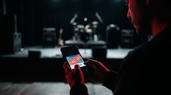 Una persona de espaldas sostiene un teléfono móvil que muestra un boleto digital con un sello rojo "CANCELADO" sobre un código de barras, con un escenario oscuro de fondo.