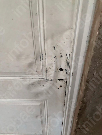 Una puerta de madera blanca entreabierta con marcas y raspones junto a la cerradura permite ver un piso de baldosas en el interior