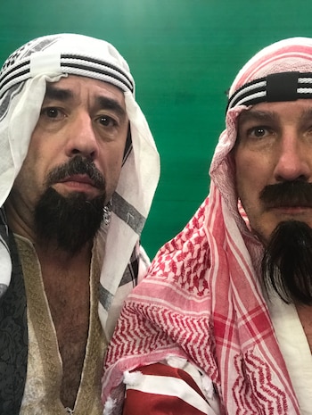 Dos hombres con pañuelos tradicionales en la cabeza y barbas postizas posan frente a un fondo verde