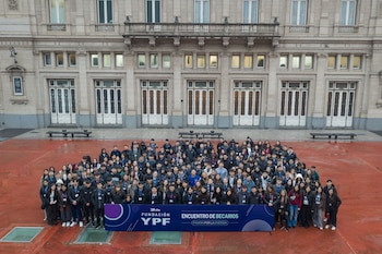 Grupo de jóvenes de pie en plaza de color rojo ladrillo frente a un edificio histórico de piedra, un cartel de la Fundación YPF en primer plano