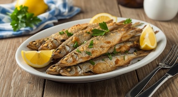 Un plato blanco contiene varios pescados pequeños dorados, gajos de limón y hojas de perejil. Un tenedor y un cuchillo están sobre una mesa de madera.
