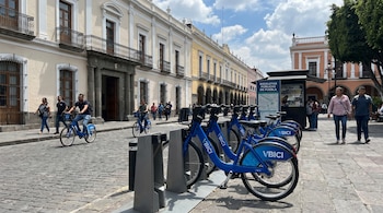 Bicicletas públicas azules "VBICI" en una estación y dos ciclistas en una calle adoquinada de Puebla. Edificios coloniales y peatones al fondo.