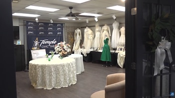 Sala de exposición con maniquíes exhibiendo vestidos, mesas con manteles decorativos, un telón azul de Temple Civic Theatre y una corona en puerta