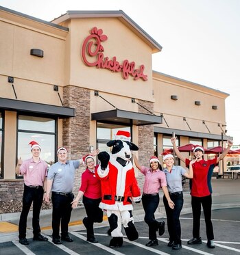 Seis empleados de Chick-fil-A y la mascota de la vaca, vestida de Papá Noel, posan frente al restaurante bajo el letrero. Todos llevan gorros de Navidad