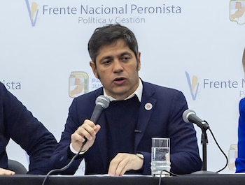 Hombre de saco oscuro y camisa blanca con un micrófono. En la mesa, un vaso de agua. Detrás, un cartel con el texto 'Frente Nacional Peronista'