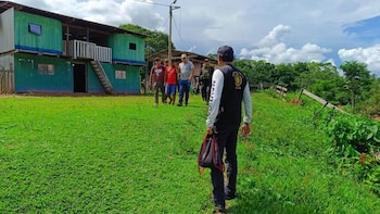 Agentes policiales y autoridades trasladan a un detenido frente a una casa verde de madera en una zona rural con vegetación