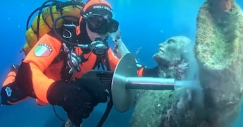 Buzo con traje de neopreno naranja y equipo de buceo limpiando una estatua sumergida. La estatua presenta crecimiento marino y desprendimiento de sedimento