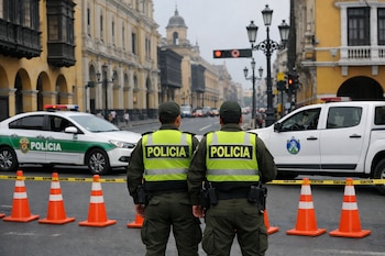 Dos policías de espaldas, patrullero, camioneta, conos naranjas, cinta amarilla, edificios coloniales, faroles y señales de tráfico en una calle.