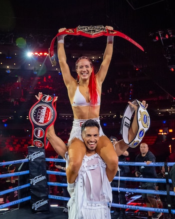 Flor Vigna, con cabello rojo y vestida de blanco, sonríe y levanta dos cinturones de campeonato, siendo cargada sobre los hombros de un hombre en un ring