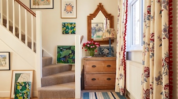 Escalera alfombrada, mueble de madera con jarrón de tulipanes, un espejo, cuadros colgados y cortinas estampadas junto a una ventana.