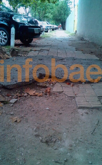 Vereda deteriorada con baldosas rotas y tierra, un tronco de árbol en el lateral y la rueda de un automóvil, con el logo de Infobae
