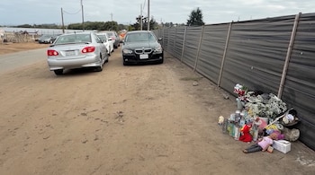 Varios vehículos estacionados junto a un cerco gris en un camino de tierra y un memorial con flores, velas y juguetes en el suelo