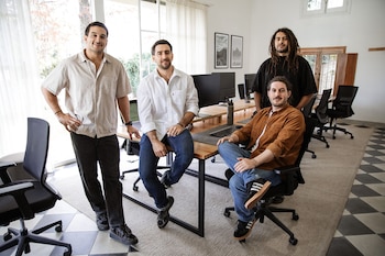 Cuatro hombres posan en una oficina moderna. Hay tres de pie y uno sentado. Se observan escritorios, computadoras, sillas y ventanas grandes