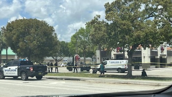 Una camioneta de policía negra, cinta amarilla de escena del crimen, personas, una furgoneta blanca y un restaurante Taco Bell bajo un cielo nublado