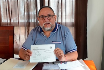 Hombre barbudo con gafas y polo azul sostiene un documento blanco del ISSSTE con escritura manual, sentado frente a cortinas marrones