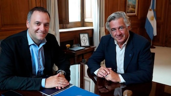 Manuel Adorni y Nicolás Pino sentados en una mesa de madera, ambos sonriendo. Al fondo, se aprecia una bandera argentina y un cuadro con un diseño enmarcado