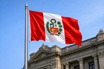 Una bandera de Perú con franjas rojas y blancas, el escudo nacional central, ondea en un mástil metálico. Se ve una fachada de edificio y cielo azul.