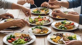 Primer plano de múltiples manos de diferentes personas sirviéndose comida de varios platos con alimentos equilibrados, incluyendo vegetales, cereales y huevos, sobre una mesa de madera.